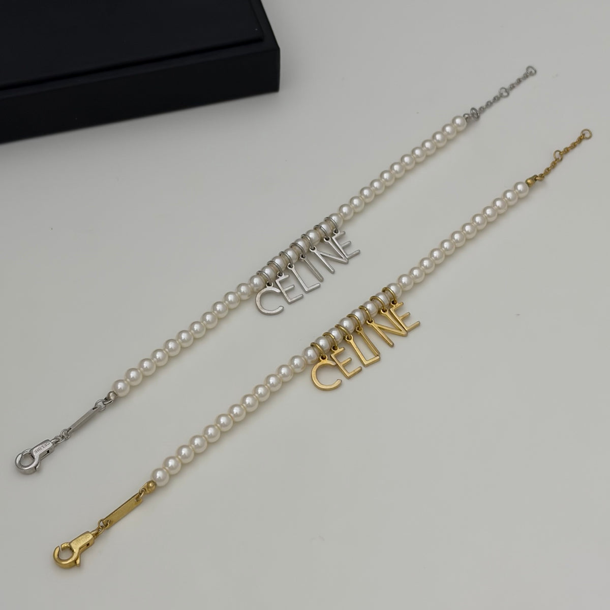 Two pearl bracelets with 'CELINE' letter charms, one in silver and one in gold, displayed on a light gray surface with a black box in the corner.