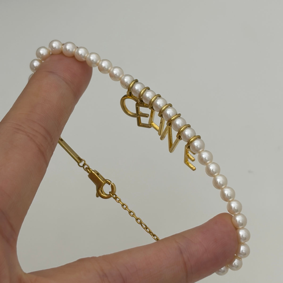 A pearl bracelet with gold lettering spelling 'CELINE' and a gold clasp, held by a person's fingers against a light gray background.