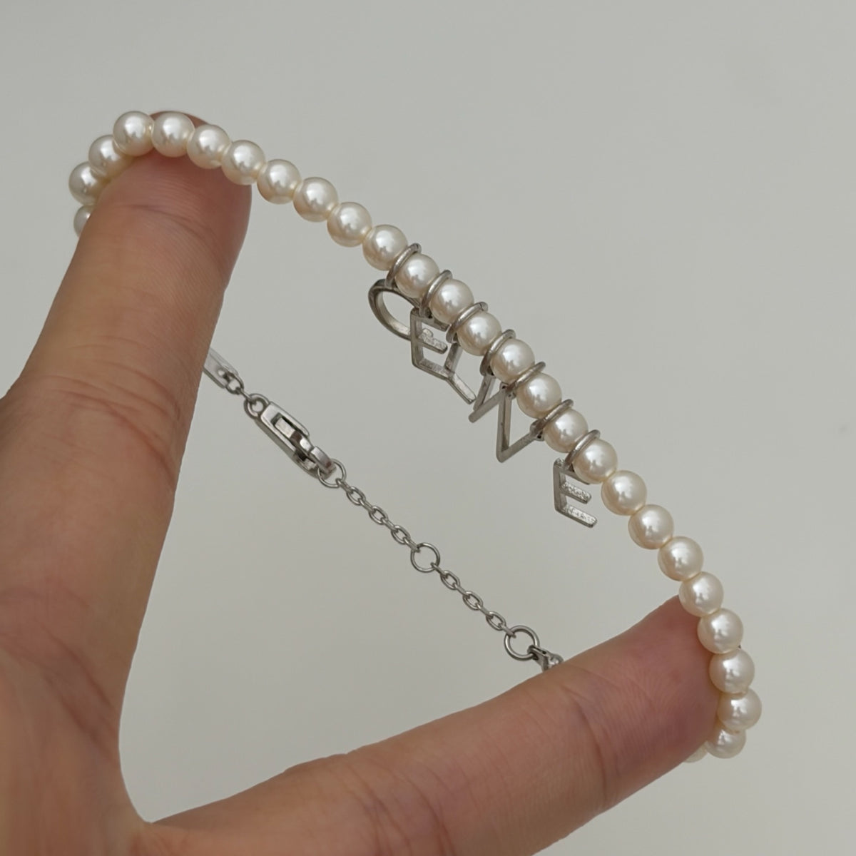 A close-up of a hand holding a pearl bracelet with silver letter charms spelling LOVE, shown against a plain light gray background.