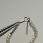 A close-up of a pearl bracelet with a silver lobster clasp and extender chain, held by metal pliers against a light gray background.