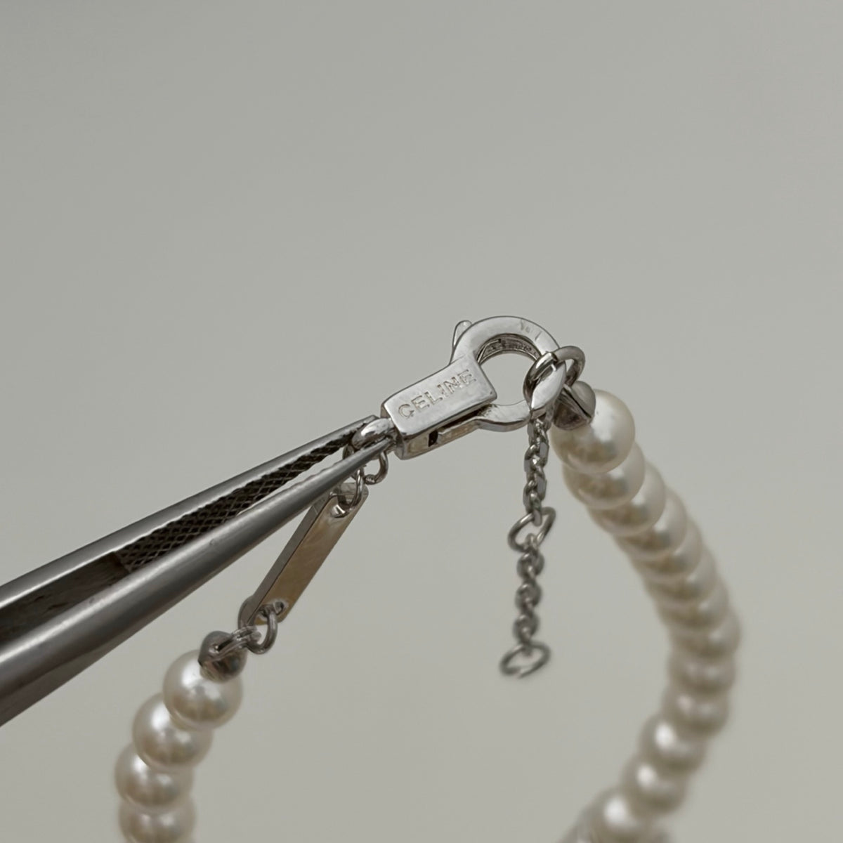 A close-up of a pearl bracelet with a silver lobster clasp and extender chain, held by metal pliers against a light gray background.