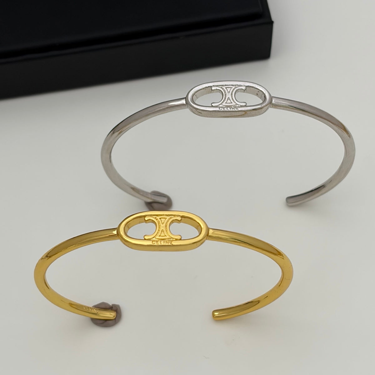 Two slim cuff bracelets with oval center motifs featuring a stylized 'H' emblem and the engraved brand name 'HÉLÈNE', shown on a light neutral background with a black box partially visible in the upper left.