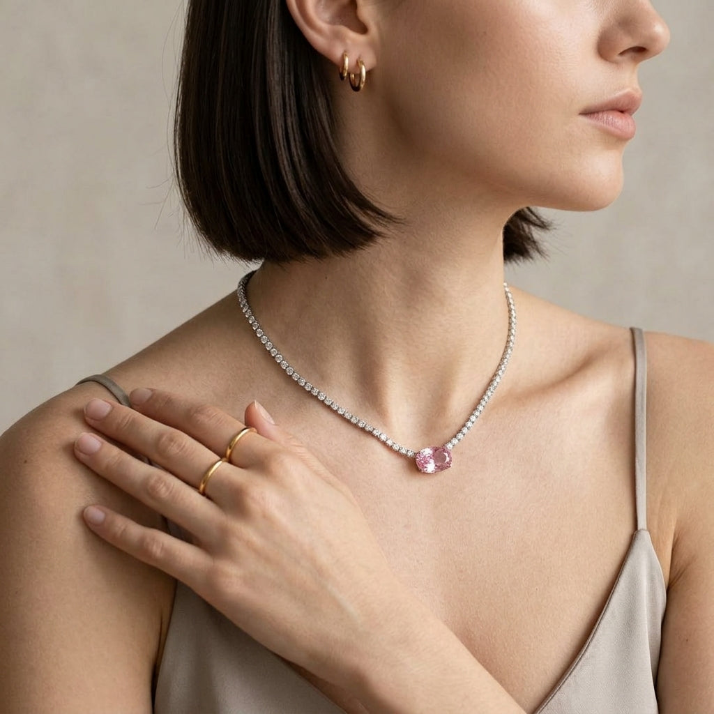 A close-up lifestyle shot of a model wearing a delicate silver-toned necklace with a round pink gemstone pendant, paired with gold rings and hoop earrings.