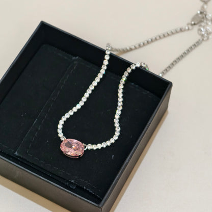 A silver-tone chain necklace with a central oval pink gemstone pendant, displayed inside a black presentation box on a light beige surface.