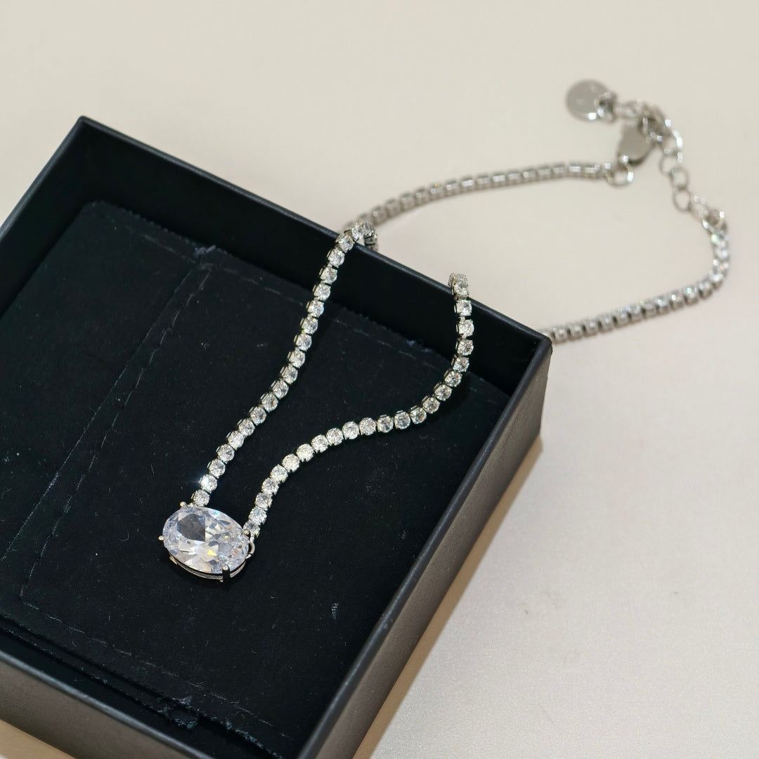 A silver-toned chain necklace with a large oval clear crystal pendant, displayed inside an open black jewelry box on a light beige surface.