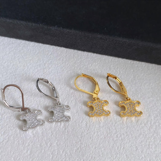 Three pairs of leverback earrings displayed on a white textured surface, including one silver pair, one gold pair, and one rose gold pair, each featuring a small shield-shaped charm paved with clear stones.