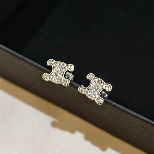 A pair of gold-toned stud earrings with a puffy, rounded silhouette covered in small clear crystals, resting on the edge of a black jewelry box.