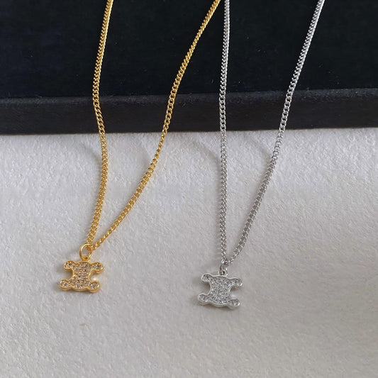 Two pendant necklaces displayed on a white textured surface with a black background. One necklace is gold-toned with a small shield-shaped pendant, and the other is silver-toned with a matching shield pendant.
