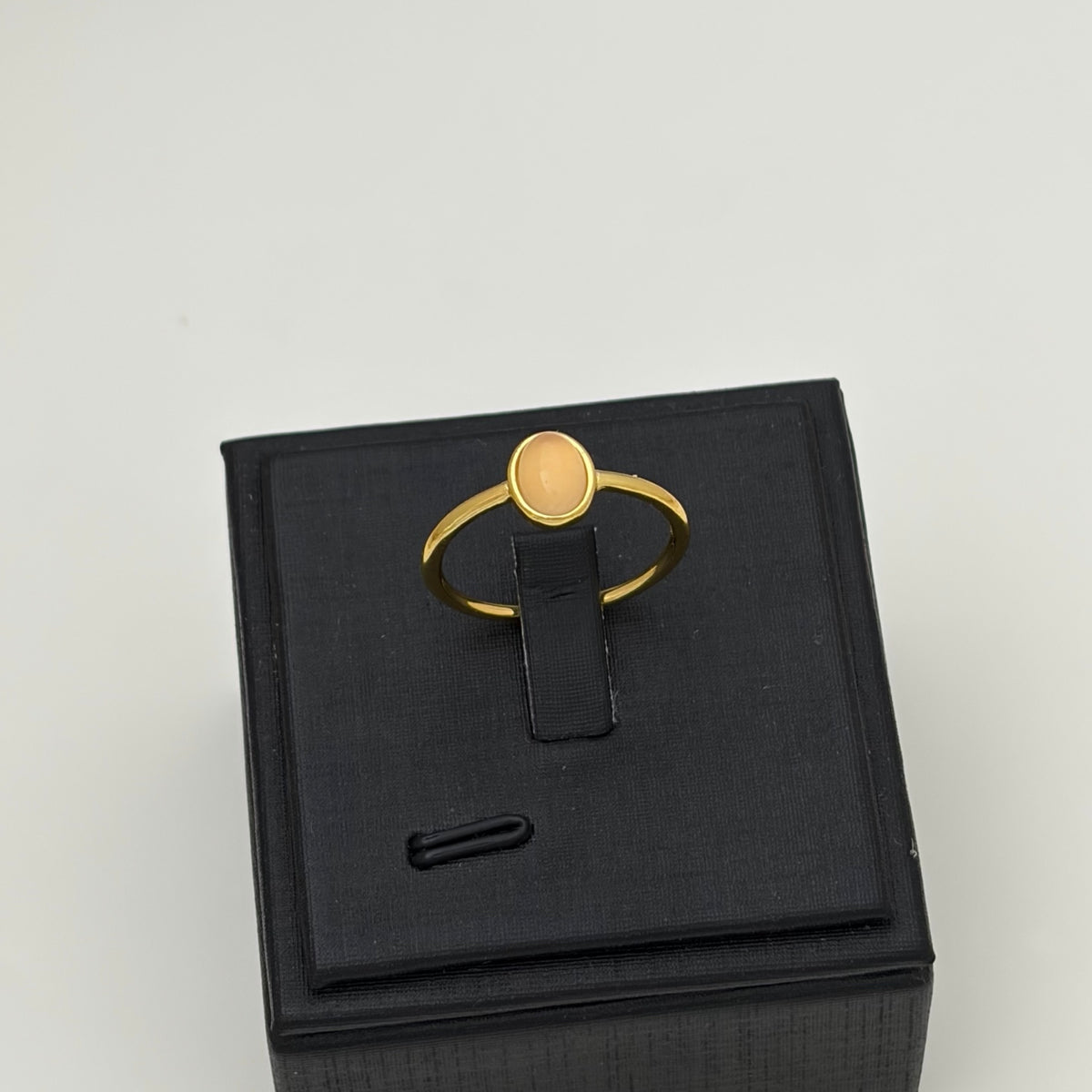 A gold ring with a round, light peach-colored stone, displayed on a black jewelry box stand against a light gray background.