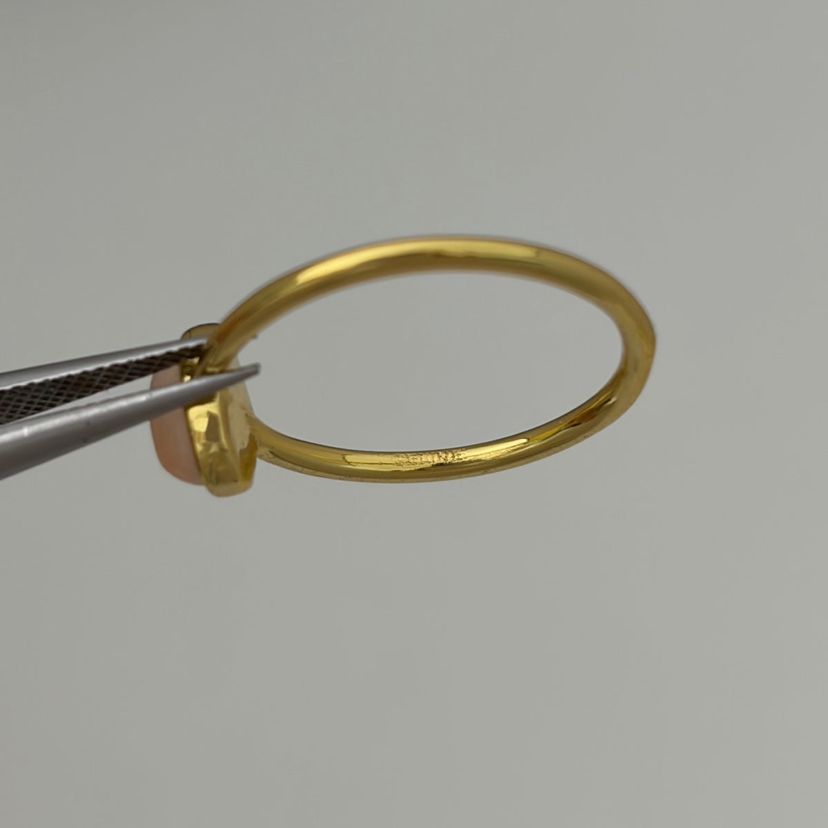 A close-up of a polished gold ring held by silver tweezers against a plain light gray background.