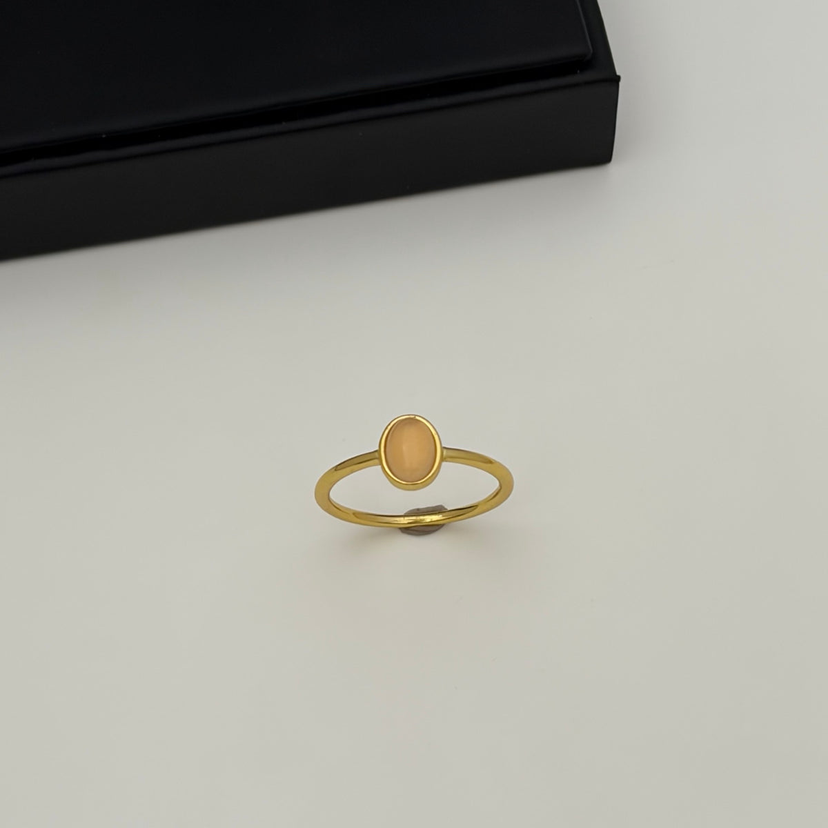 A gold ring with a small oval peach-colored stone, displayed on a light gray surface with a black box in the background.