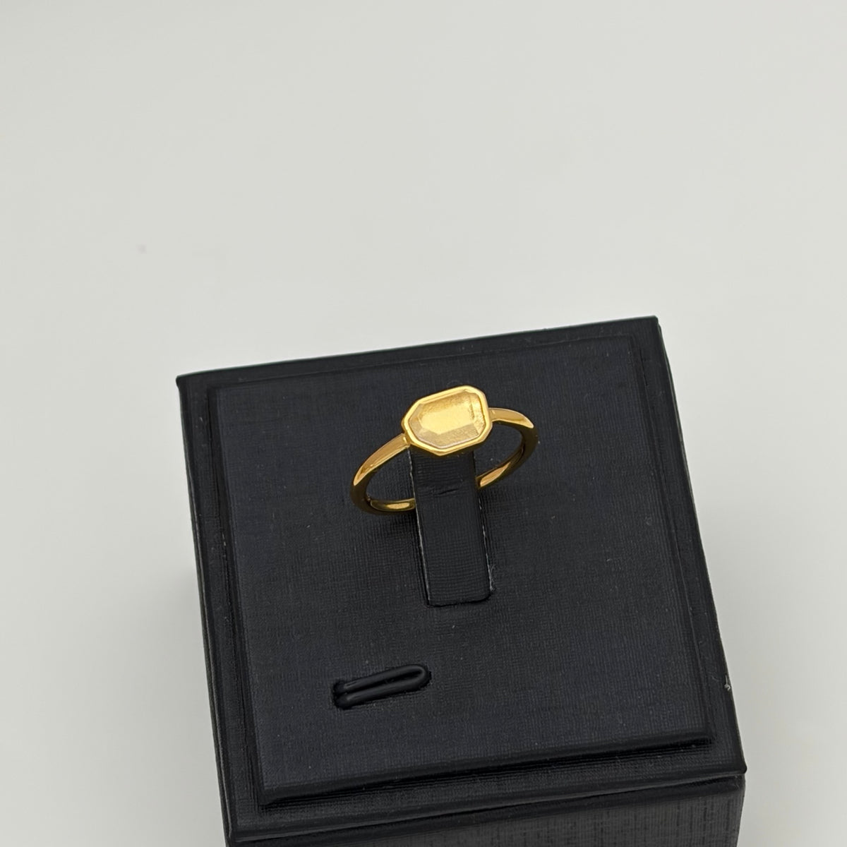 A gold signet ring with a rectangular bezel-set stone, displayed on a black jewelry box against a light gray background.