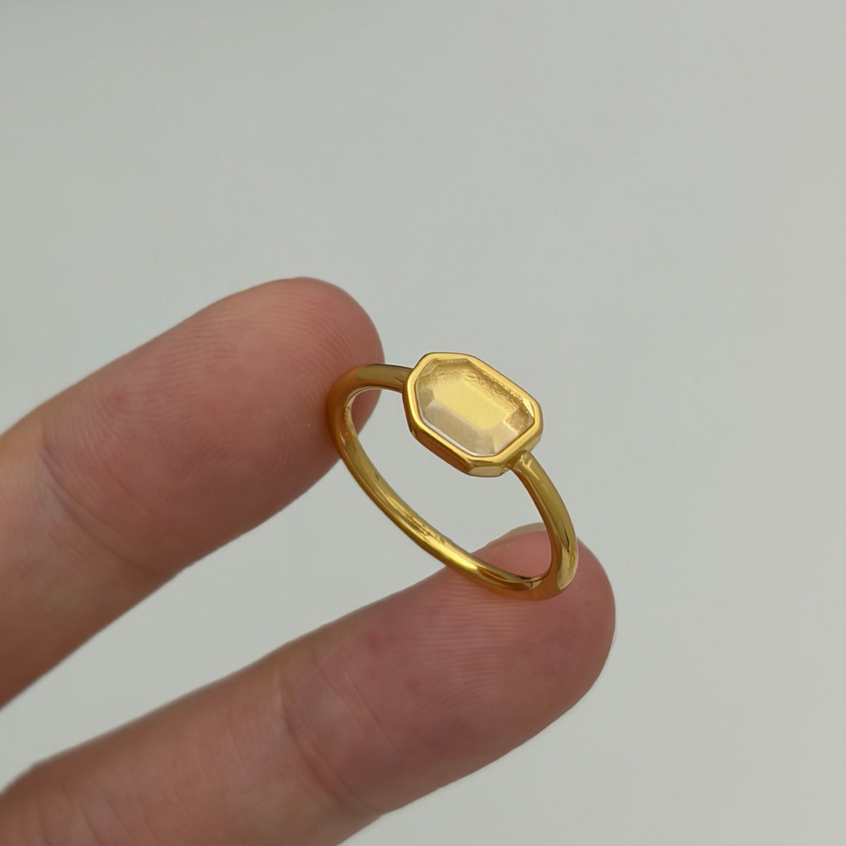 A gold ring with a faceted, beveled clear gemstone held between two fingers against a plain light gray background.
