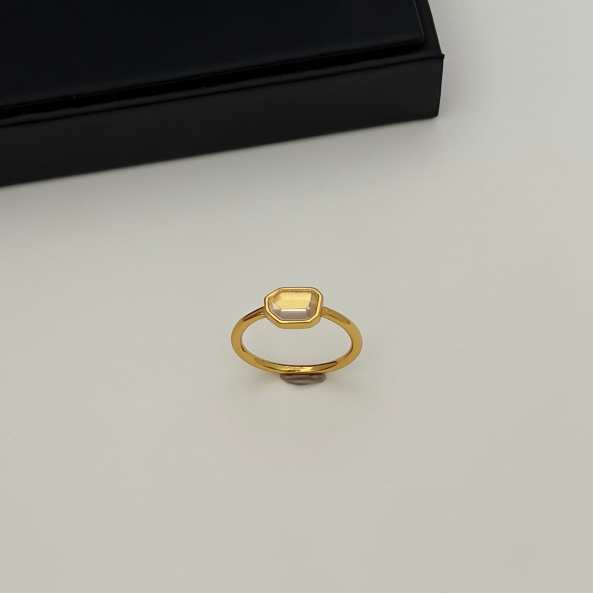 A gold ring with a small, rounded rectangular yellow gemstone, displayed on a light gray surface with a black box in the background.