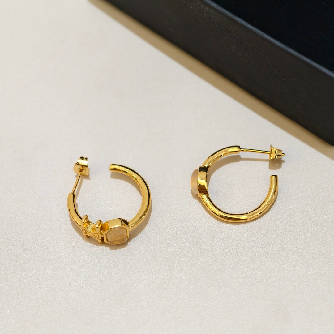 A pair of gold hoop earrings with a small geometric charm on the front, displayed on a light beige surface next to a black box.