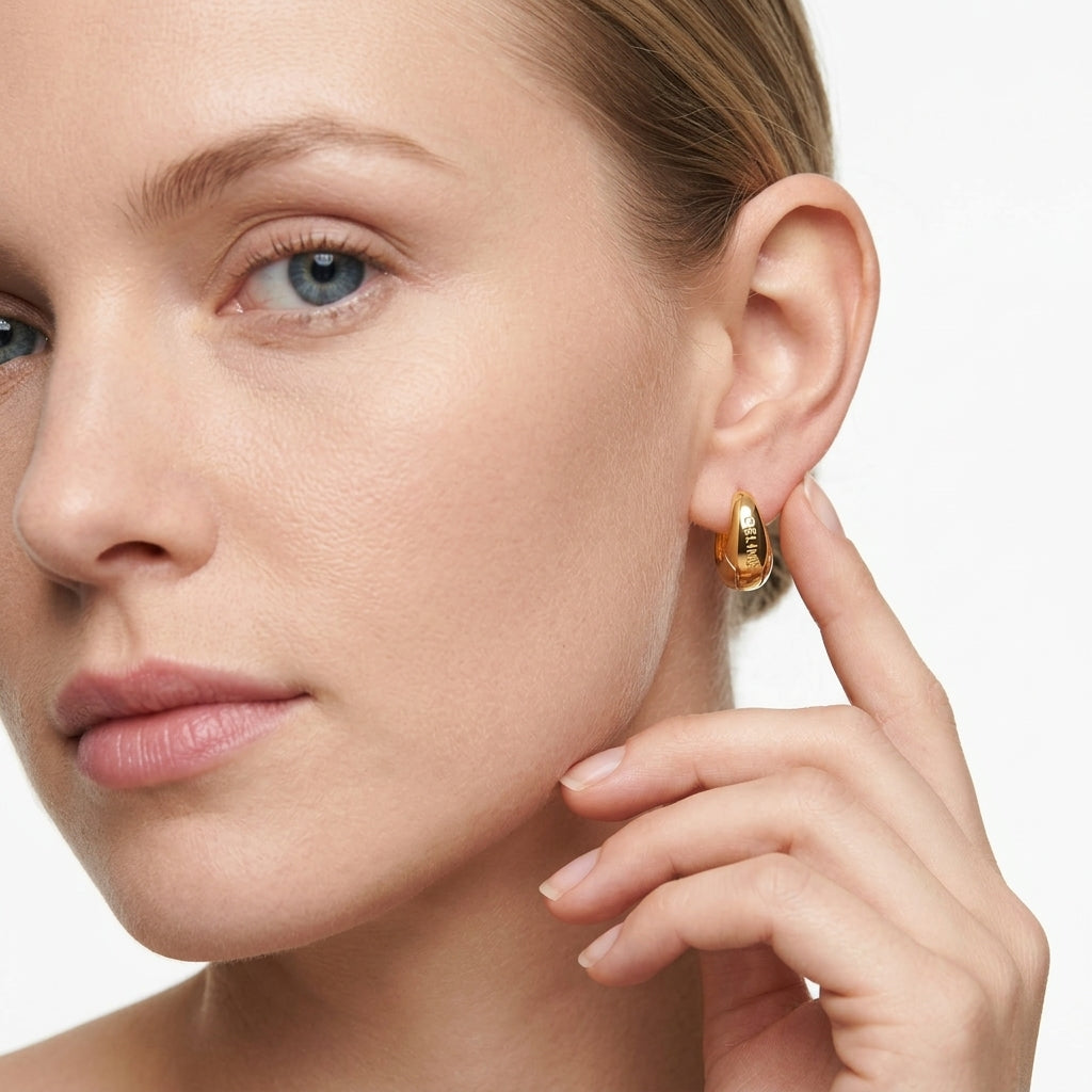 A close-up of a woman's face and ear wearing a small gold hoop earring, with her hand near her neck against a clean white background.