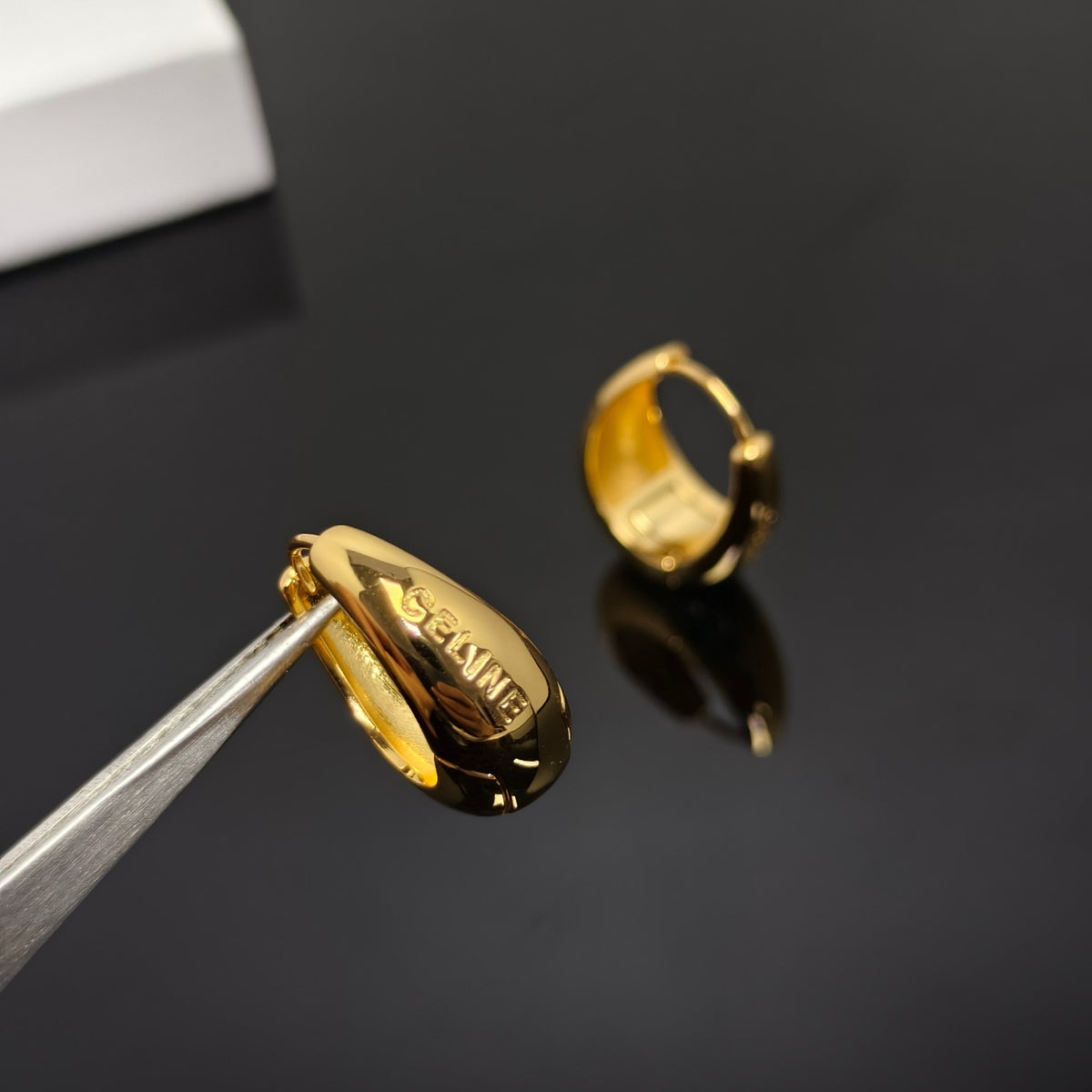 A gold huggie hoop earring held by metal tweezers, with the matching earring ring standing on a glossy black reflective surface in the background.