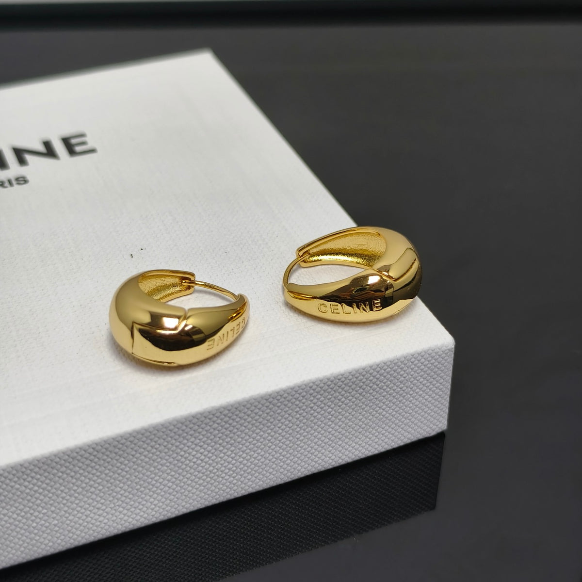 A pair of gold hoop earrings with a chunky, organic curved shape, resting on a white textured Celine box. The brand name 'CELINE' is engraved on the side of the earrings, and 'PARIS' is partially visible on the box.