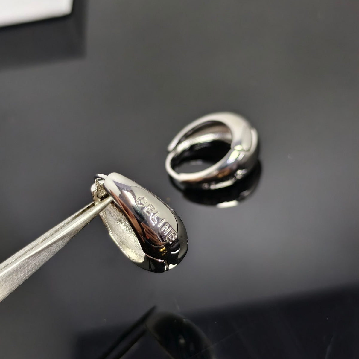 A polished silver huggie hoop earring with a teardrop-shaped front, held by metal tweezers in the foreground, with the matching earring resting in the background on a glossy black surface. The front of the earring is engraved with the brand name 'CELINE'.