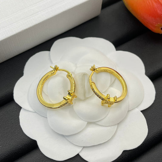 A pair of gold hoop earrings displayed on a white flower-shaped stand, with a white box in the background and a blurred orange object in the top right corner.