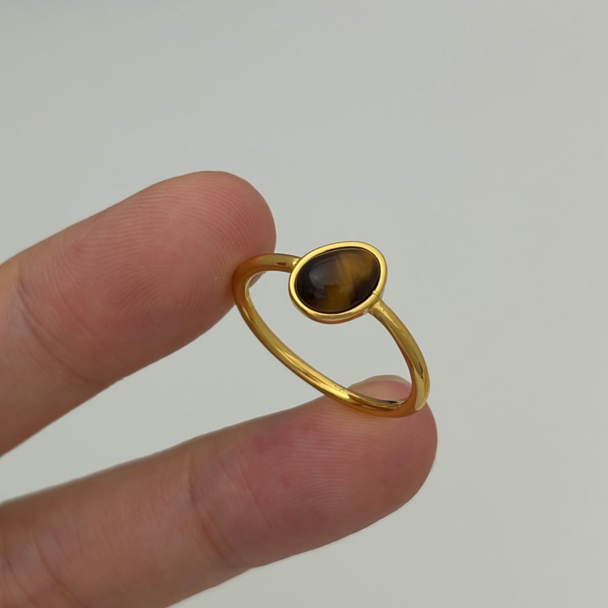 A gold ring with an oval tiger's eye stone held between two fingers against a plain light gray background.