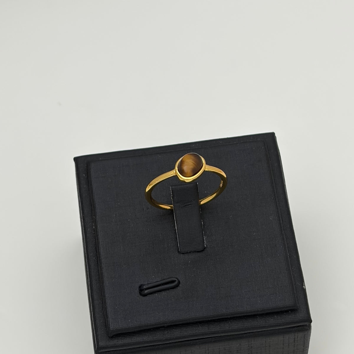 A gold ring with a round brown cabochon gemstone, displayed on a black jewelry box stand against a light gray background.