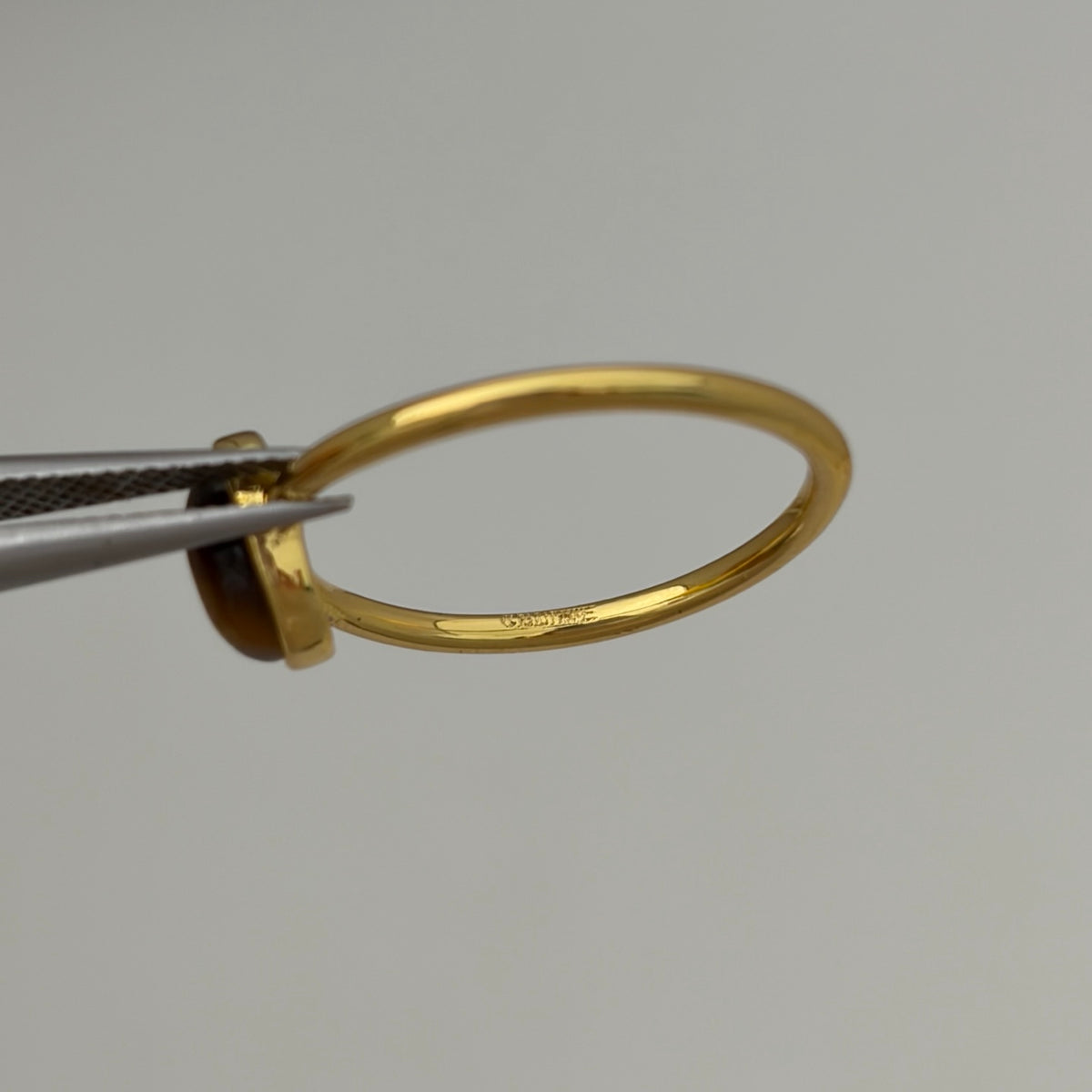 A close-up of a polished gold ring held by silver tweezers against a plain light gray background.