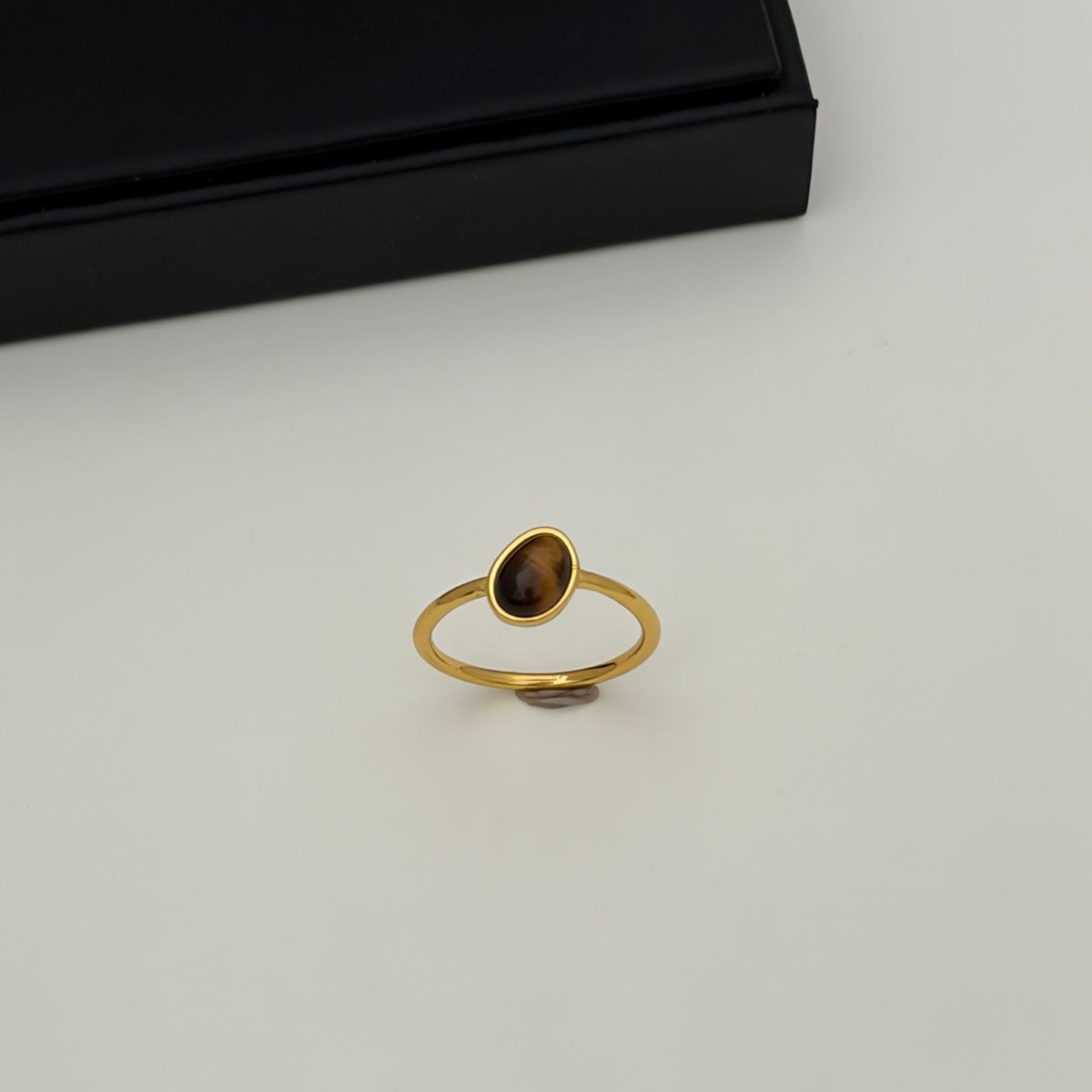 A gold ring with a small, teardrop-shaped brown gemstone, displayed on a light gray surface with a black presentation box in the background.