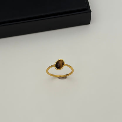 A gold ring with a small, teardrop-shaped brown gemstone, displayed on a light gray surface with a black presentation box in the background.