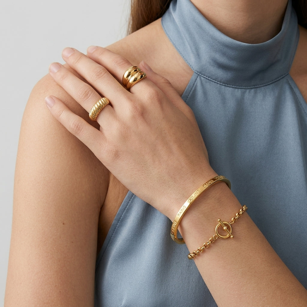 A close-up of a model wearing gold jewelry, including a textured bangle bracelet on the wrist, a ring on the middle finger, and a stacked ring on the index finger, wearing a dusty blue high-neck top against a light gray background.