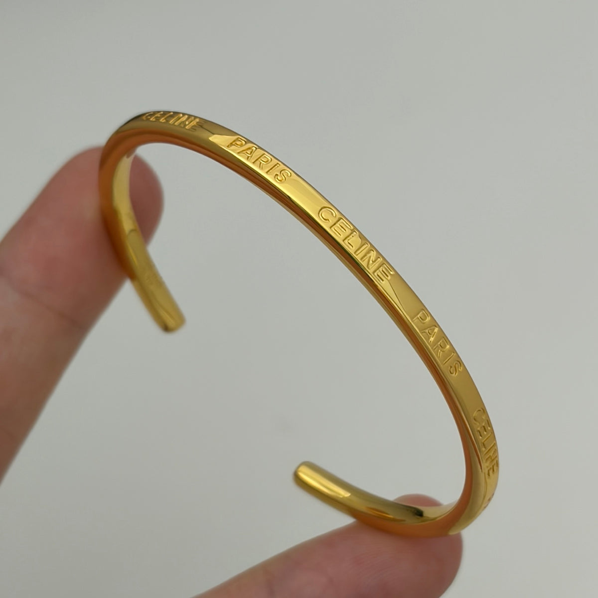 A gold-tone Celine cuff bracelet engraved with 'CELINE PARIS' text, held by a person's fingers against a plain light gray background.