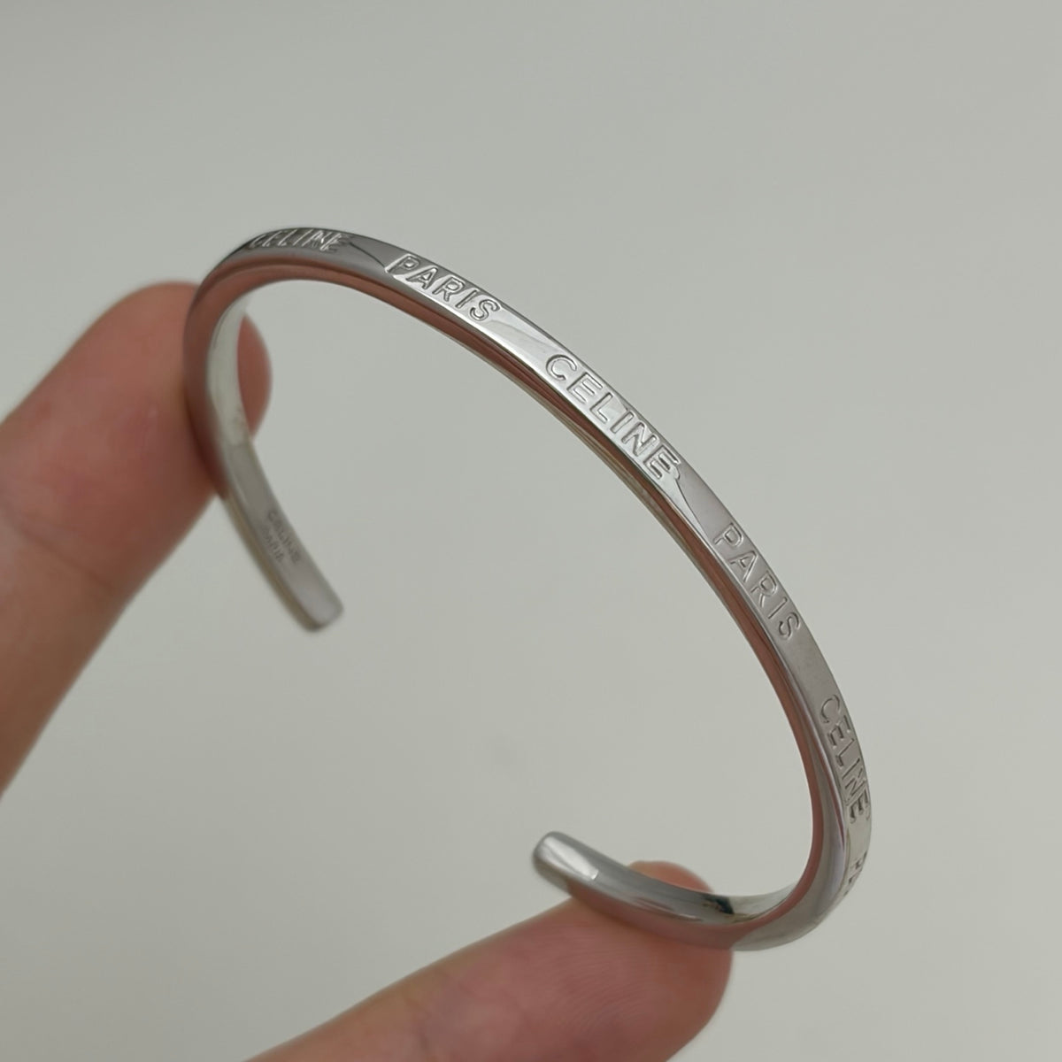 A silver-toned metal cuff bracelet engraved with 'PARIS CÉLINE' and 'MONT LUX' being held by a person's fingers against a plain light gray background.