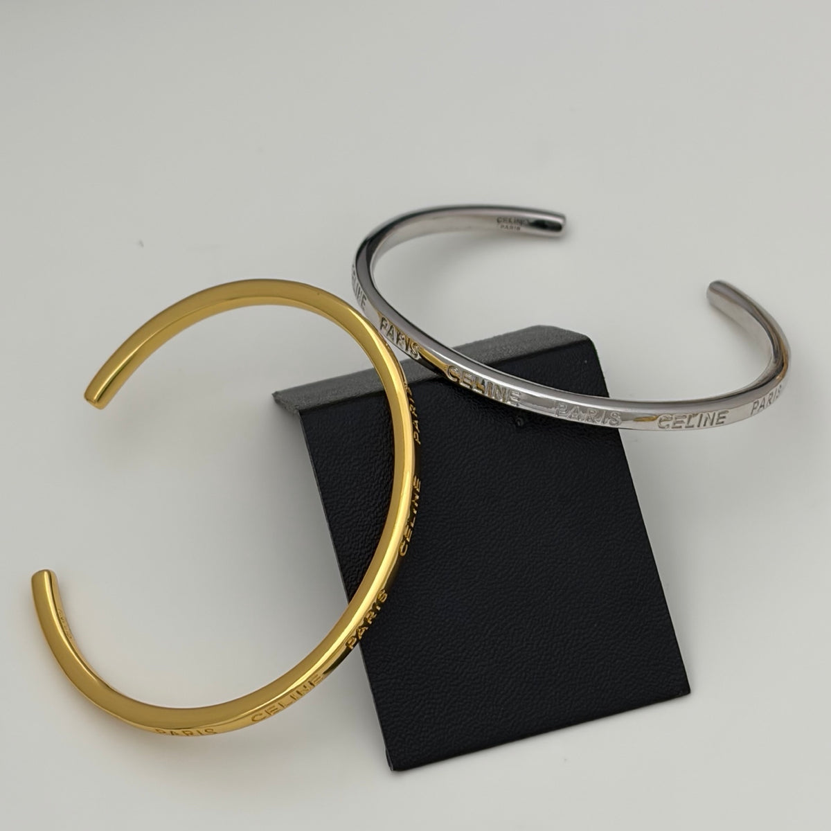 Two slim open cuff bracelets, one gold-toned and one silver-toned, both engraved with 'CELINE PARIS', displayed on a black rectangular stand against a light gray background.
