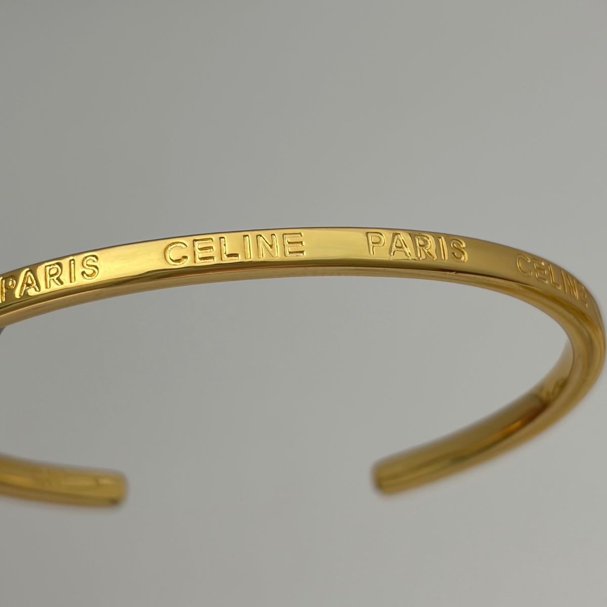 A gold-tone open cuff bangle bracelet engraved with 'PARIS CELINE' repeatedly, shown against a neutral light gray background.