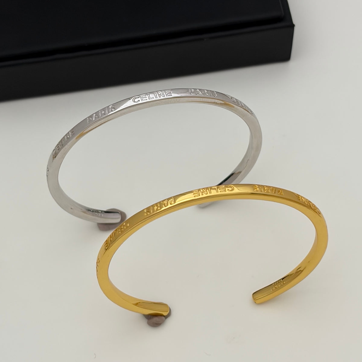 Two open cuff bracelets, one silver-toned and one gold-toned, both engraved with the name 'MARA', displayed on a light surface with a black box in the background.