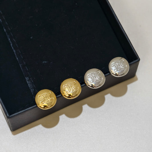 Four round metal studs with an embossed monogram design, shown in gold and silver finishes resting on a black jewelry box against a light gray background.