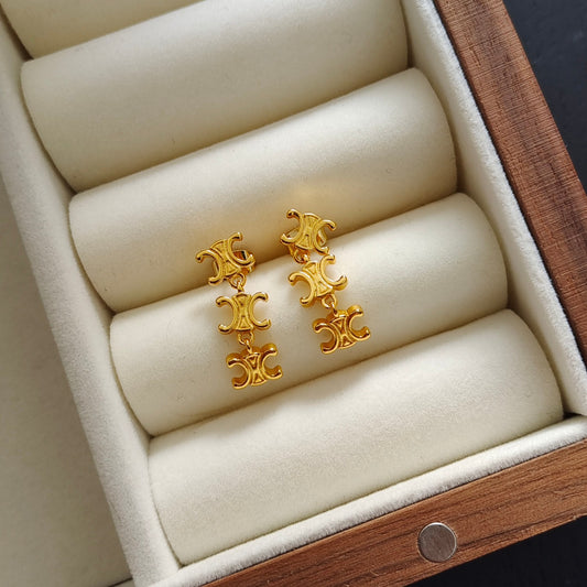 A pair of gold drop earrings with stacked ornate Celine-style triple-C logos, displayed on cream velvet jewelry rolls inside a wooden presentation box.