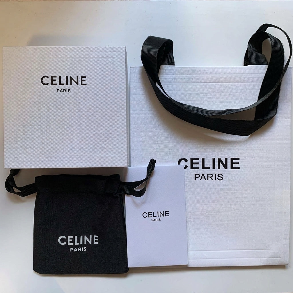 Celine Paris branded packaging set including a white textured box, white shopping bag with black handles, black dust bag, and a white card on a white background.