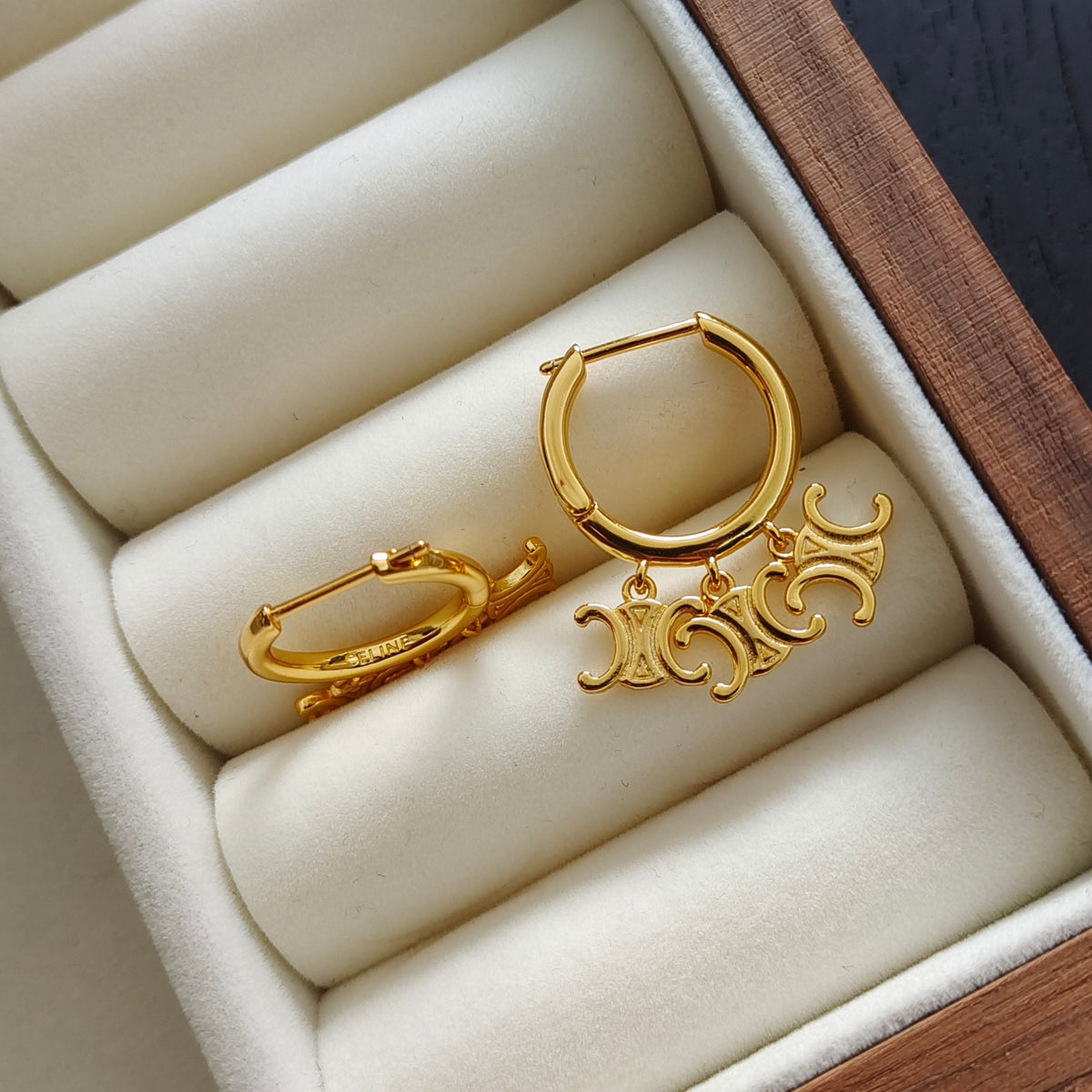 A pair of gold hoop earrings with matching curved charm pendants, displayed on cream-colored cushioned rings inside a wooden jewelry box.