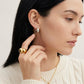 A close-up of a woman wearing a small gold hoop earring with a clear gemstone and a chunky gold ring, shown against a clean light background.