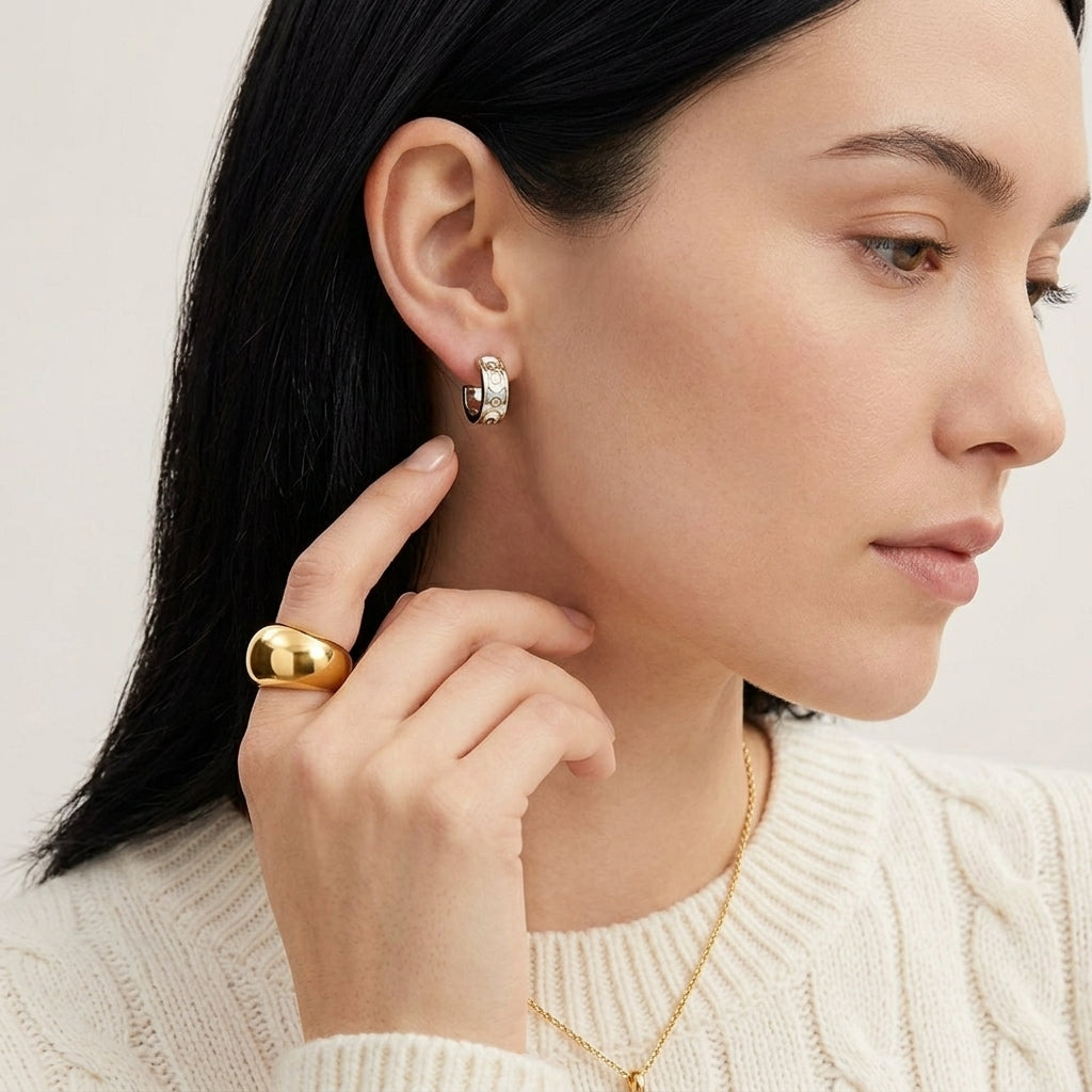 A close-up of a woman wearing a small gold hoop earring with a clear gemstone and a chunky gold ring, shown against a clean light background.