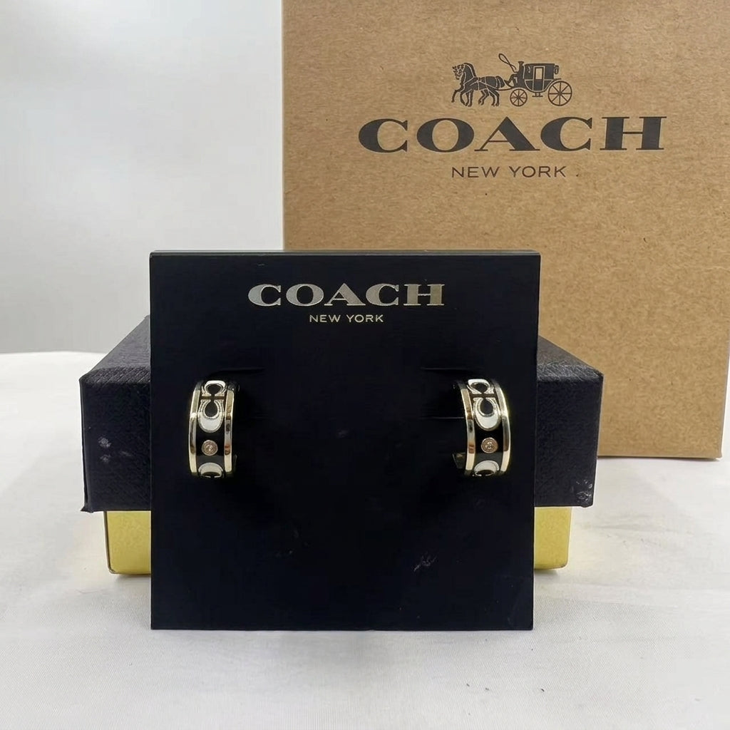 A pair of Coach New York hoop earrings displayed on a black branded card, with the Coach New York box behind them on a white surface.