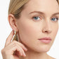 A close-up of a woman wearing a small gold huggie hoop earring with a white textured inlay, touching her ear with her hand against a clean white background.