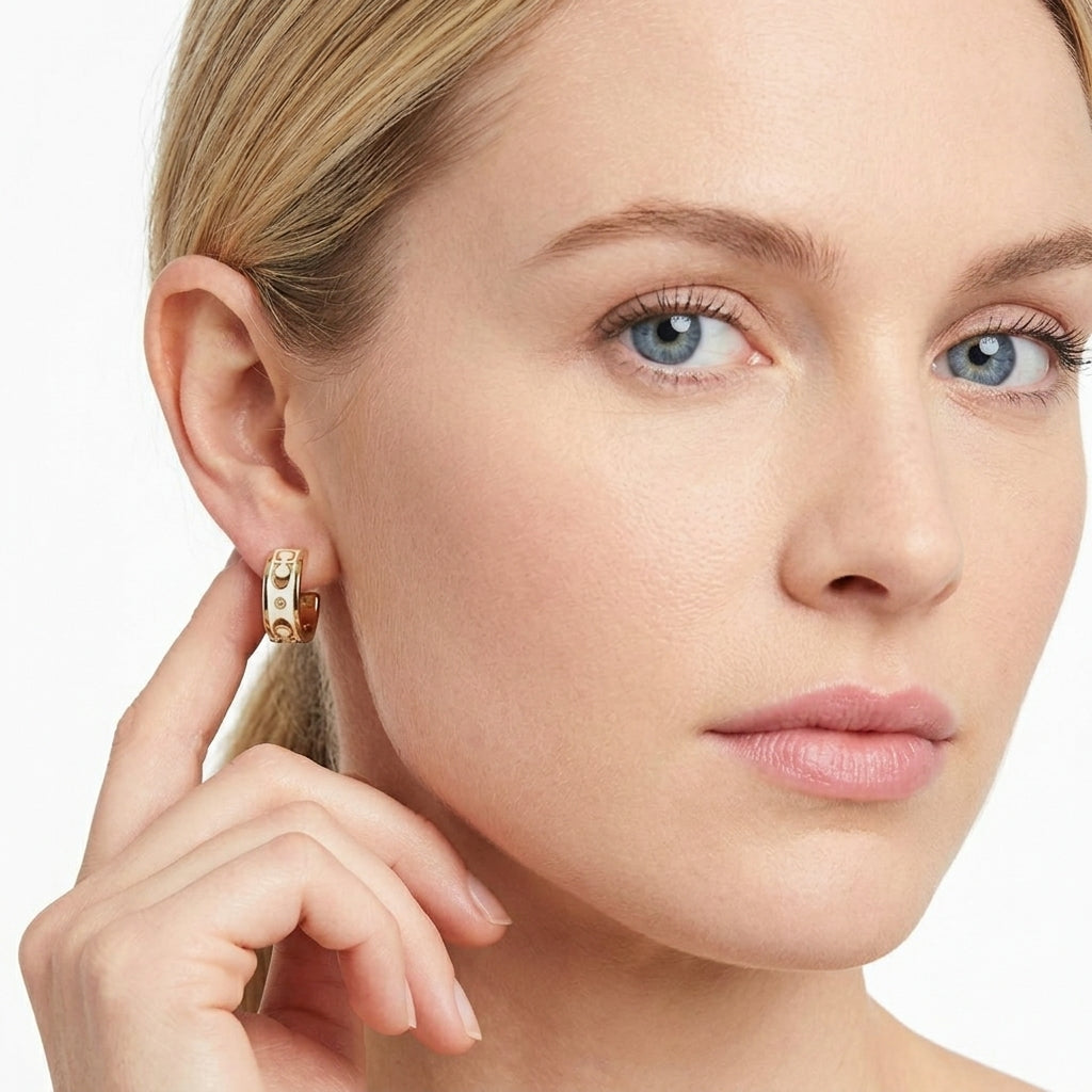 A close-up of a woman wearing a small gold huggie hoop earring with a white textured inlay, touching her ear with her hand against a clean white background.