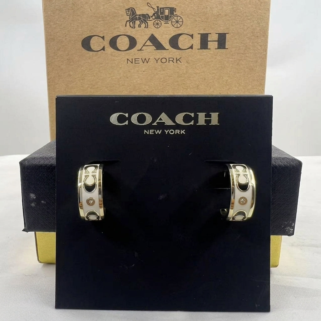 A pair of Coach New York hoop earrings displayed on a black branded card, with the kraft paper COACH box behind them on a white background.