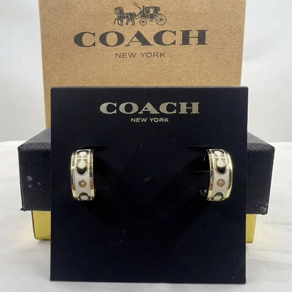 A pair of Coach New York hoop earrings displayed on a black branded card, with the kraft paper COACH box behind them on a white background.