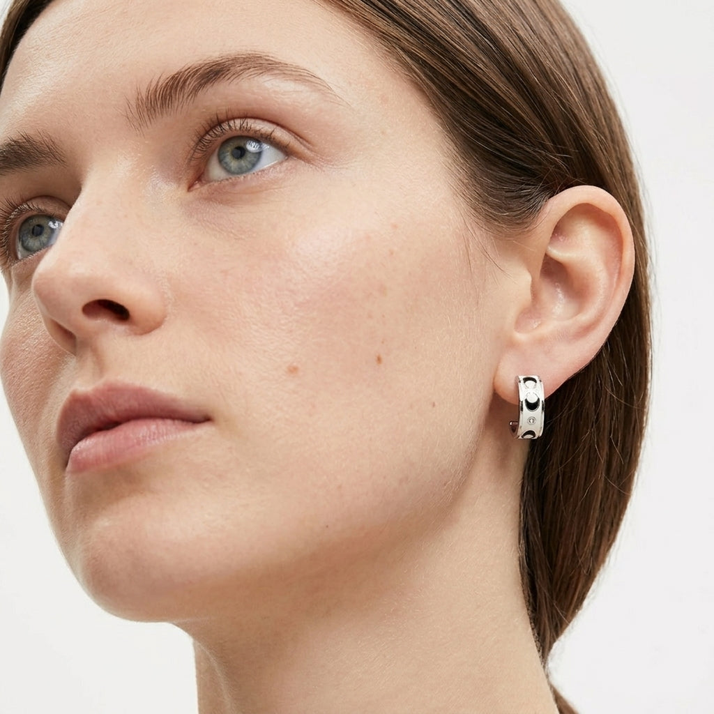 A close-up of a woman's ear wearing a small silver hoop earring with a black and white geometric pattern, set against a clean white background.