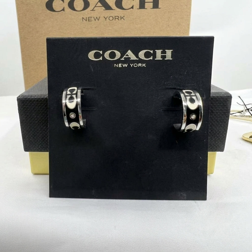 A pair of silver-tone Coach hoop earrings displayed on a black branded card, with a Coach box behind them on a white surface.