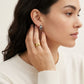 A close-up lifestyle shot of a woman wearing a small gold hoop earring and a gold ring, shown in profile against a clean off-white background.