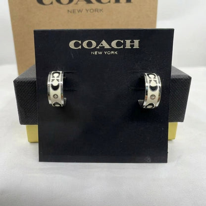 A pair of Coach huggie hoop earrings with silver-tone metal and black-and-white geometric patterns, displayed on a black Coach New York card against a brown Coach box.